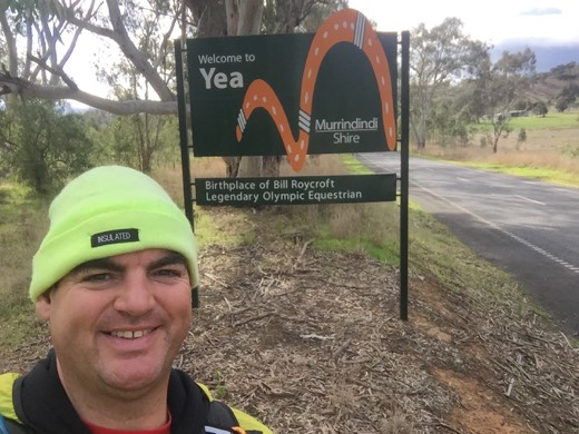 Gary smiling beside the Welcome to Yea sign during Day 84